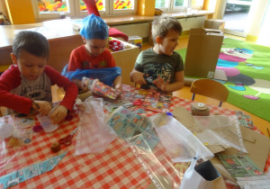 Trzech chłopców przygotowuje elementy strojów ekologicznych z papierowych ulotek, tną, kleją. Jeden z chłopców ma założoną czapkę z reklamówki na głowie.